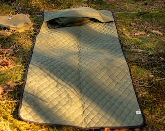 Può includere: Un tappetino da esterno trapuntato verde oliva con un bordo blu scuro è steso sul pavimento della foresta. Un cuscino verde oliva abbinato è in cima al tappetino. Una piccola borsa verde oliva è a sinistra del tappetino.