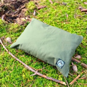 Peut inclure: Un oreiller rectangulaire vert olive repose sur un lit de mousse verte, de brindilles et de pommes de pin. Un petit patch noir et blanc est cousu sur l'oreiller. L'oreiller est conçu pour une utilisation en extérieur.