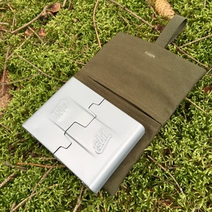 Peut inclure: Un réchaud de camping pliable en argent avec le texte "Esbit" dessus, dans un étui de transport en toile verte avec une sangle.
