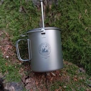 Puede incluir: Una taza de camping de titanio plateado con asa y tapa, con la inscripción "NORDIC WOLF GERMANY TITANIUM 750ml". Incluye una prensa francesa. La taza está sobre musgo verde.