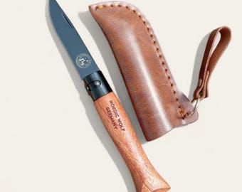 May include: A black folding knife with a wooden handle and a leather sheath. The knife has a black blade and a black handle with the words "Nordic Wolf Germany" engraved on it.