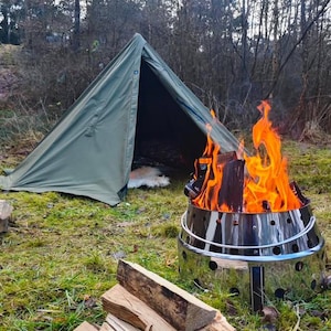Könnte beinhalten: Ein grünes Campingzelt, das in einem Wald aufgebaut ist, mit einer Feuerstelle davor. Die Feuerstelle ist aus Metall und hat ein Feuer, das darin brennt. Neben der Feuerstelle liegen Holzscheite gestapelt.