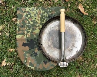Puede incluir: Una sartén redonda de metal con un mango de madera y una bolsa de tela de camuflaje. La sartén se utiliza para cocinar sobre fuego abierto.