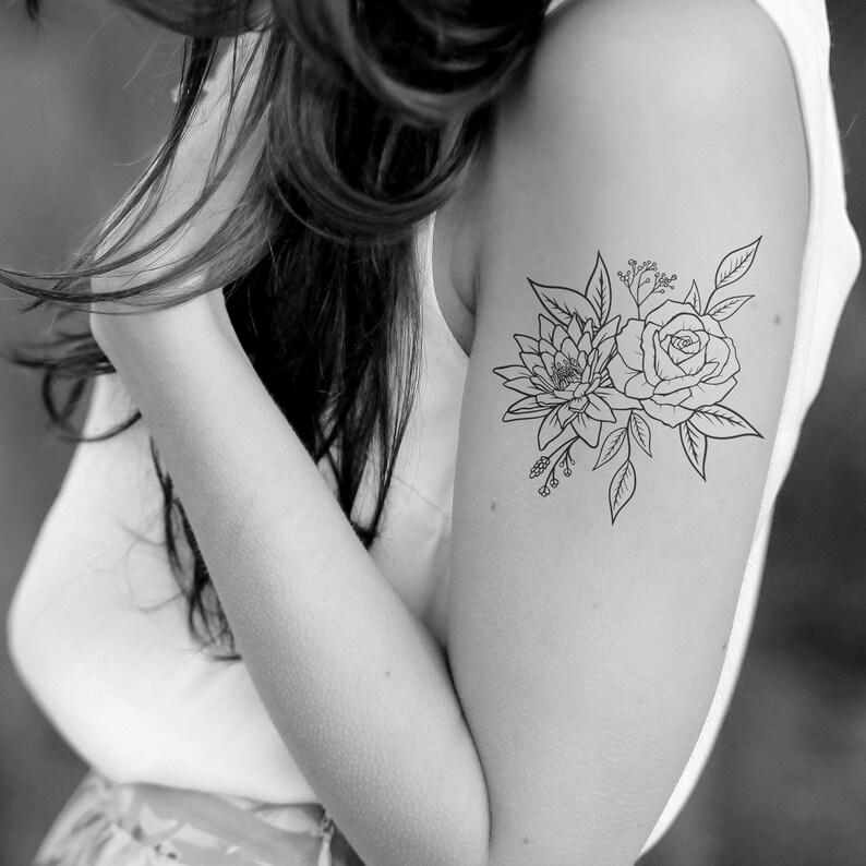 May include: Black and white floral temporary tattoo featuring a rose and a dahlia with leaves and sprigs. The tattoo is applied to the upper arm. The design is detailed and artistic, suitable for body art.