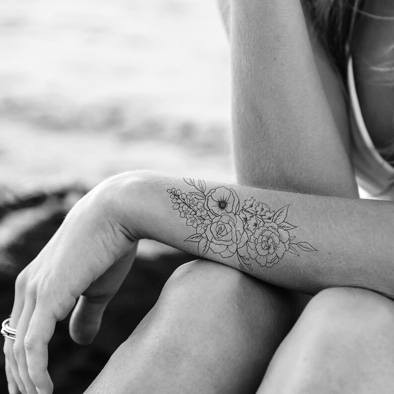 May include: Black and white floral temporary tattoo applied to the forearm. The tattoo features a bouquet of roses, peonies, and other flowers with detailed line art. The design is a delicate and elegant floral arrangement.