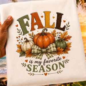 May include: A white t-shirt featuring a graphic with the text FALL is my favourite SEASON in green, orange, and brown. The design includes three pumpkins in orange, white, and green, accented by autumn leaves, berries, and a green plaid bow.