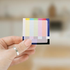 May include: A color chart with a variety of colored squares, including yellow, pink, blue, and green. The chart is held in a hand with a silver ring.