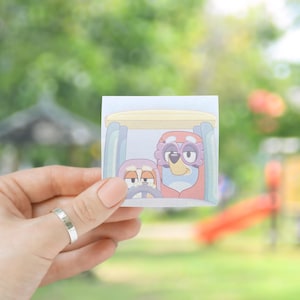 May include: A close-up of a paper note with a cartoon illustration of two animals driving a small vehicle. The animals are purple and blue with large eyes and are wearing sunglasses. The vehicle is yellow and has a white roof.