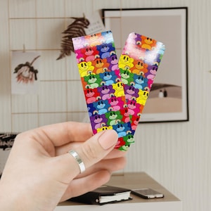 May include: Two rainbow-colored bookmark strips with a repeating pattern of smiling cartoon bears. The bears are arranged in rows and are outlined in black. The bookmarks are made of a shiny, reflective material.