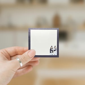 May include: A blank white note card with a black border and a cartoon dog peeking out from the bottom right corner. The dog is gray and white with black eyes and a black nose.
