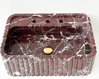 May include: A rectangular red marble sink with a white veining pattern. The sink has a fluted edge and a gold drain stopper.