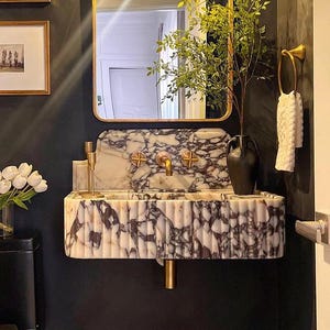 Op de afbeelding: Een witte marmeren wastafel met een gouden kraan en decoratieve accenten. De wastafel is gemonteerd op een zwarte muur en heeft een uniek, geribbeld ontwerp.