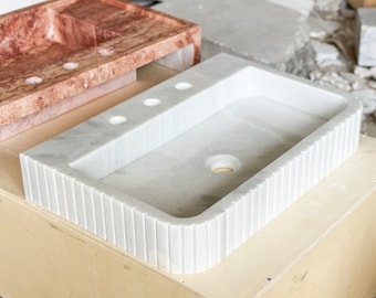 Op de afbeelding: Een witte marmeren wastafel met een rechthoekige vorm en een afgeronde rand. De wastafel heeft een afvoergat en drie kleine gaten aan de bovenkant. De wastafel staat op een houten oppervlak.