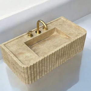 Op de afbeelding: Een beige stenen badkamer wastafel met een rechthoekige vorm en een geflute rand. De wastafel heeft een enkele kraan met twee handvatten, alles in een gouden afwerking.