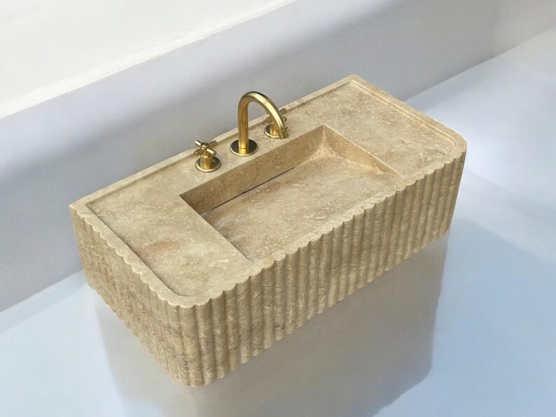 Op de afbeelding: Een beige stenen badkamer wastafel met een rechthoekige vorm en een geflute rand. De wastafel heeft een enkele kraan met twee handvatten, alles in een gouden afwerking.