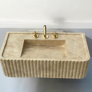 Op de afbeelding: Een beige stenen badkamer wastafel met een rechthoekig bassin en een gegroefde rand. De wastafel heeft drie gouden kranen.