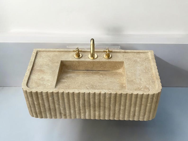 Op de afbeelding: Een beige stenen badkamer wastafel met een rechthoekig bassin en een gegroefde rand. De wastafel heeft drie gouden kranen.