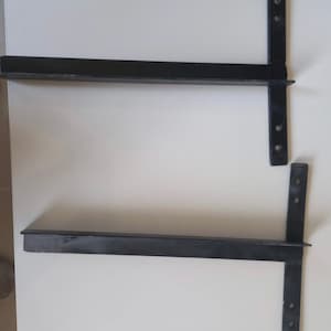 May include: Two black metal shelf brackets with multiple holes for mounting. The brackets are L-shaped and have a flat surface for attaching a shelf.