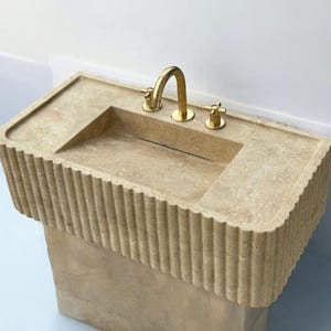 Op de afbeelding: Een beige stenen badkamer wastafel met een gouden kraan en twee knoppen. De wastafel staat op een sokkel met een geribbeld ontwerp.
