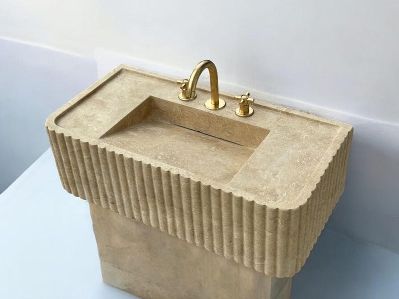 Op de afbeelding: Een beige stenen badkamer wastafel met een gouden kraan en twee knoppen. De wastafel staat op een sokkel met een geribbeld ontwerp.