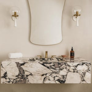 Op de afbeelding: Een moderne badkamer met een marmeren wastafelblad en geïntegreerde wastafel. De wastafel heeft goudkleurige kranen en een bijpassende spiegel. Twee wandlampen met glazen bollen flankeren de spiegel. Een opgerolde witte handdoek en een zeepdispenser staan op het aanrecht.