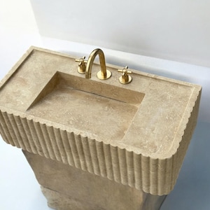 Op de afbeelding: Een beige stenen badkamer wastafel met een rechthoekig bassin en een gouden kraan. De wastafel heeft een decoratieve, gegroefde rand en staat op een voet.