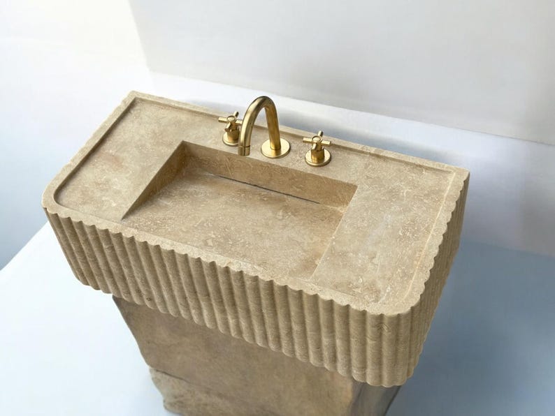 Op de afbeelding: Een beige stenen badkamer wastafel met een rechthoekig bassin en een gouden kraan. De wastafel heeft een decoratieve, gegroefde rand en staat op een voet.