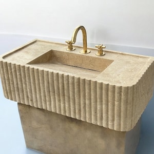 Op de afbeelding: Een beige stenen badkamer wastafel met een rechthoekig bassin en een gouden kraan. De wastafel heeft een decoratief, geribbeld ontwerp rond de rand.