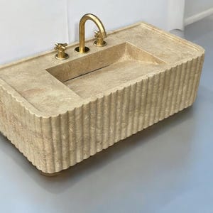 Op de afbeelding: Een beige stenen badkamer wastafel met een geribbelde rand en drie gouden kranen.