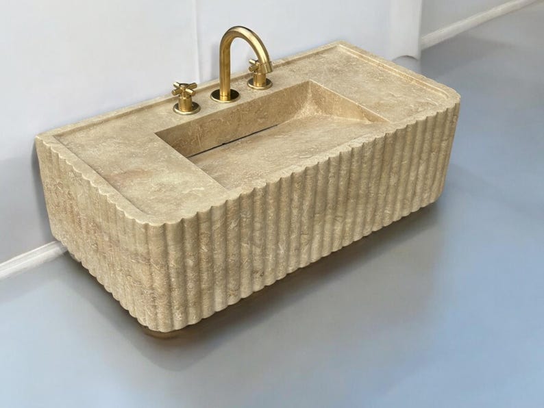 Op de afbeelding: Een beige stenen badkamer wastafel met een geribbelde rand en drie gouden kranen.