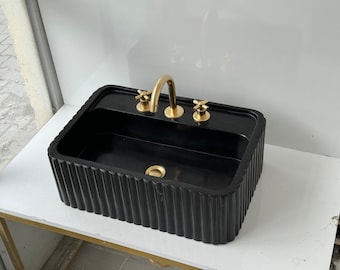 Könnte beinhalten: Schwarzes rechteckiges Badezimmerwaschbecken mit einem goldenen Wasserhahn und zwei goldenen Griffen. Das Waschbecken hat ein geripptes Design an den Rändern.