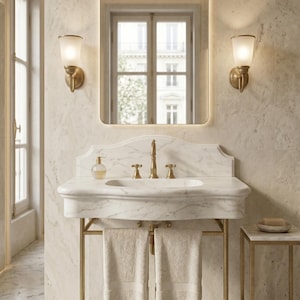 May include: A white marble bathroom vanity with a brass metal frame, featuring a matching marble backsplash, a brass faucet, and two hanging beige towels. Above the vanity is a rectangular mirror with rounded corners and two brass wall sconces.