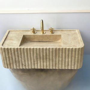 Op de afbeelding: Een beige stenen badkamer wastafel met een rechthoekig bassin en een geflute basis. De wastafel heeft drie gouden kranen.