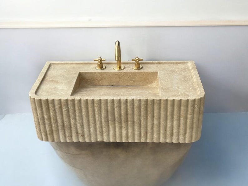Op de afbeelding: Een beige stenen badkamer wastafel met een rechthoekig bassin en een geflute basis. De wastafel heeft drie gouden kranen.