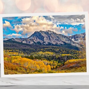 May include: A scenic mountain landscape with a peak covered in snow and a valley filled with vibrant yellow, orange, and green trees. The sky is blue with white clouds.