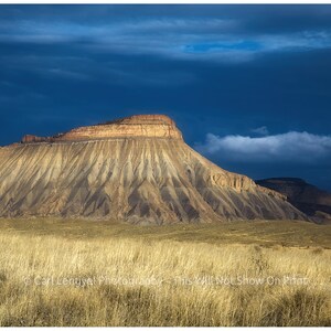 May include: A large, layered rock formation with a dark blue sky and white clouds in the background. The rock is a mix of brown, yellow, and gray colors. The foreground is a field of dry, brown grass.