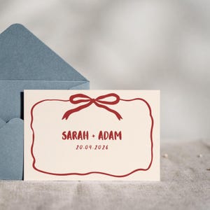 Puede incluir: Una tarjeta de invitación crema con un lazo y borde rojos, con los nombres "SARAH + ADAM" y la fecha "20.04.2026". La tarjeta está frente a un sobre azul, ideal para una boda o evento especial.