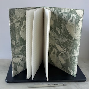 May include: Open notebook with lined pages, resting on a laptop. The cover features a green and white pelican design. A silver pen with the text "ElegantWriter" is in the foreground. The notebook is open, displaying several pages.