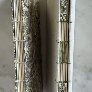 May include: Two hand-bound books with exposed stitching on the spines. The left book features a marbled cover in shades of green, blue, and cream. The right book has a plain cream fabric cover with green thread and white pearl bead accents.