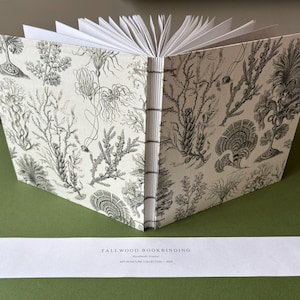May include: A journal with a cream-colored cover featuring black botanical illustrations of seaweed and coral. The book has an exposed stitched spine and white pages. A white paper strip below reads: TALLWOOD BOOKBINDING, Handmade Journal, ART IN NATURE COLLECTION - 2024.