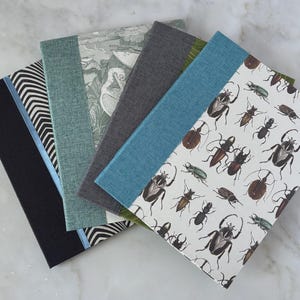 May include: A collection of notebooks with various cover designs. The covers include a black and white striped pattern, a botanical illustration, and a white background with insect illustrations. The notebooks are arranged in a fan shape.