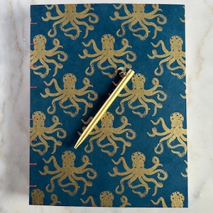 May include: A teal notebook with a gold octopus pattern and red stitching. A gold pen rests diagonally across the notebook. The notebook's cover features a repeating design of stylized octopuses.