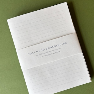 May include: A stack of white, pre-folded lined bookbinding signatures held by a paper band labeled TALLWOOD BOOKBINDING, Pre-Folded Lined Bookbinding Signatures, 6 x 8 in., Short Grain, Ready to Sew. The paper is white with thin gray lines.
