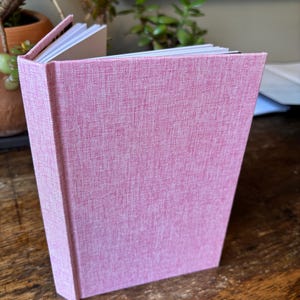 May include: A hardcover book with a textured, dusty rose pink fabric cover. The book is standing upright and slightly open, revealing white pages along the top edge. The fabric material has a visible cross-hatch weave pattern throughout the surface.