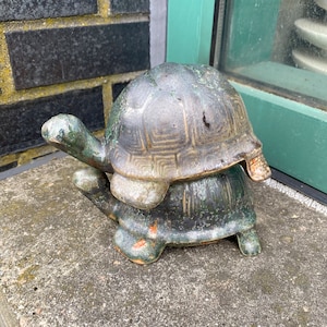 May include: A ceramic turtle sculpture with a green and brown glaze. The turtle is stacked on top of another turtle, creating a layered effect.