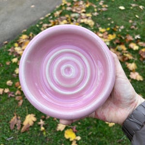May include: A pink ceramic bowl with a spiral pattern in the centre. The bowl has a glossy finish and a raised rim. The bowl is held in a hand, with a background of green grass and fallen leaves.