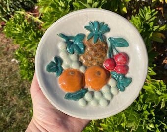 May include: A white ceramic plate with a raised design of fruit. The design includes oranges, grapes, and a pineapple. The fruit is painted in shades of orange, green, and brown.