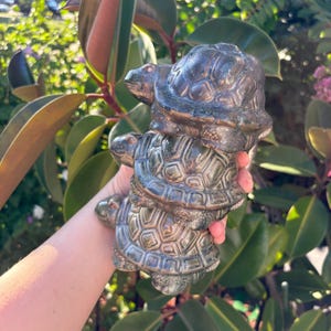 Peut inclure: Une sculpture de couleur bronze représentant trois tortues empilées les unes sur les autres. Les tortues ont des motifs de carapace détaillés. La sculpture est tenue dans une main.