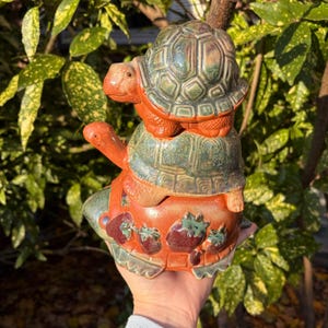 Peut inclure: Une pile de trois figurines de tortues en céramique. La tortue du bas est orange avec un glaçage vert et a des fraises sur sa carapace. La tortue du milieu est verte avec un glaçage brun et la tortue du haut est verte avec un glaçage brun.