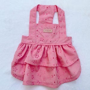 May include: A pink dog dress with eyelet detailing and a ruffled skirt. The dress has adjustable straps and a small tag near the top. The fabric has a floral pattern and is made of a lightweight material.